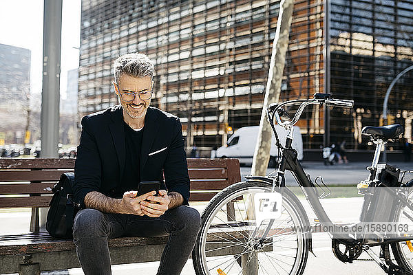 Gray-haired businessman sitting on a bench next to bicycle in the city using cell phone