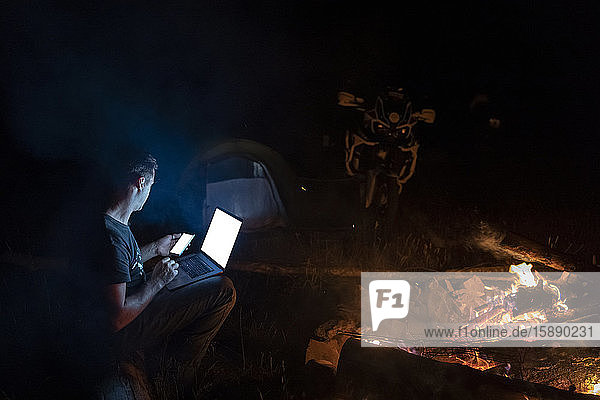 Mann  der nachts mit Laptop und Smartphone am Kamin sitzt