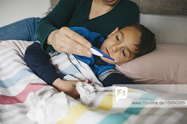 Kranker Junge mit Gewebe in der Hand liegt im Bett und schaut zusammen mit seiner Mutter auf das Digitalthermometer
