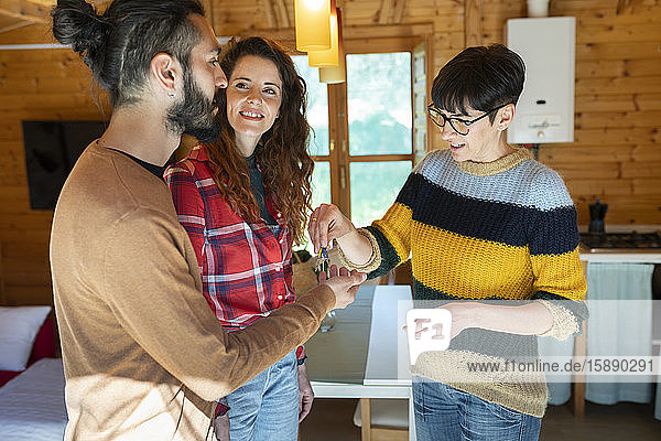 Gastgeberin empfängt junges Paar in einer Hütte auf dem Land und übergibt ihm den Hausschlüssel