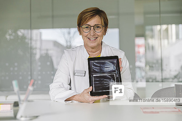 Am Schreibtisch sitzender Arzt zeigt Röntgenbild der Hand auf digitalem Tablett