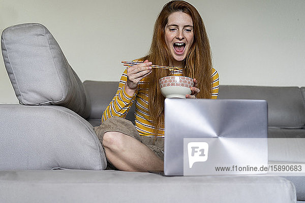 Glückliche junge Frau  die zu Hause auf der Couch sitzt und isst  während sie einen Laptop benutzt