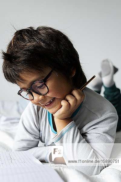 Portrait of little boy lying on bed doing homework