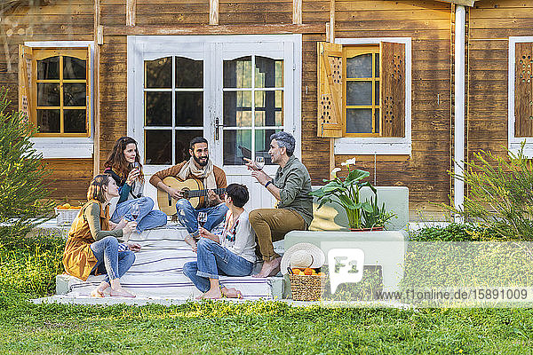 Friends playing music on the guitar and drinking wine outside a cabin in the countryside