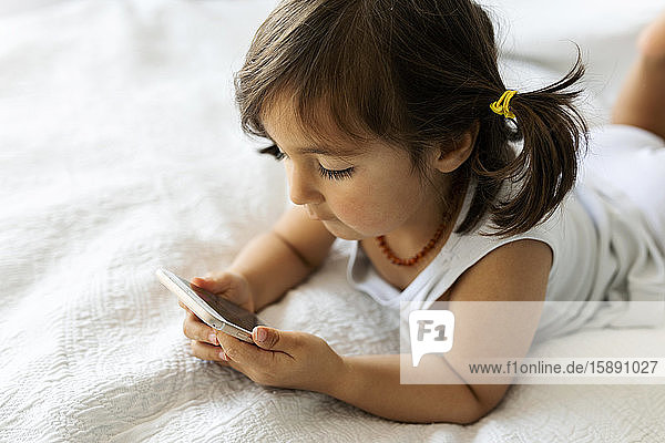 Kleines Mädchen in Unterwäsche liegt auf dem Bett und schaut auf Smartphone