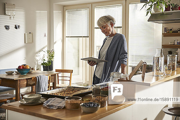 Ältere Frau steht in Küche mit digitaler Tablette bei der Zubereitung von Müsli