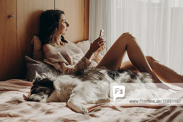 Lächelnde Frau  die mit ihrem Hund auf dem Bett liegt und aus dem Fenster schaut
