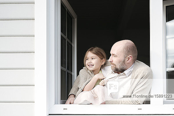 Reifer Mann mit lächelnder Tochter  die aus dem Fenster ihres Hauses schauen