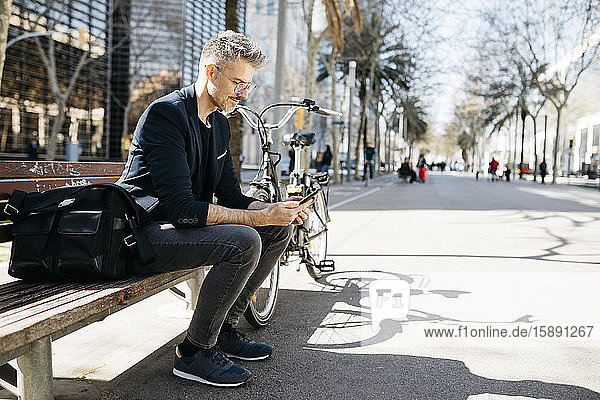 Grauhaariger Geschäftsmann sitzt in der Stadt auf einer Bank neben dem Fahrrad und benutzt sein Handy