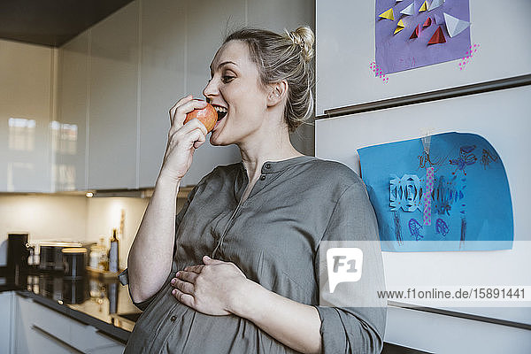 Schwangere Frau isst zu Hause in der Küche einen Apfel