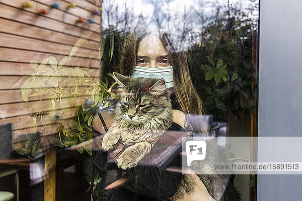 Porträt eines Mädchens mit chirurgischer Maske und Katze hinter Fensterscheibe