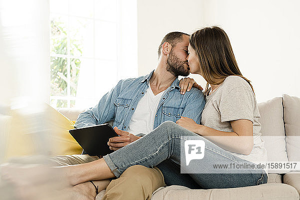 Affectionate couple relaxing at home and kissing while sitting on couch and holding tablet