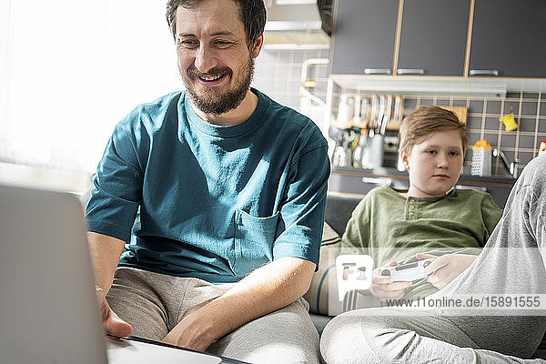 Porträt eines lächelnden Vaters  der zu Hause einen Laptop benutzt  während sein Sohn im Hintergrund langweilig ist