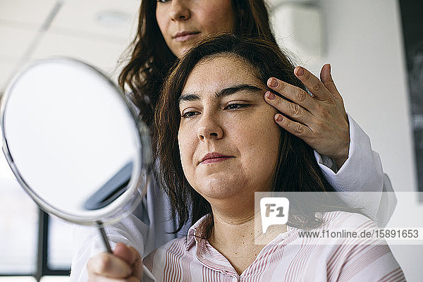 Kosmetischer Chirurg hält Spiegel und spricht mit Patient in medizinischer Praxis