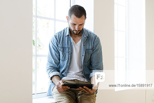 Young man at home looking at tablet sitting on window bench