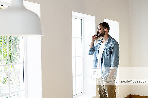 Junger Mann zu Hause im Wohnzimmer  der mit seinem Smartphone spricht  während er aus dem Fenster schaut