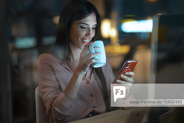 Lächelnde junge Frau mit Kaffeetasse sitzt am Schreibtisch im Büro und schaut auf ein Smartphone