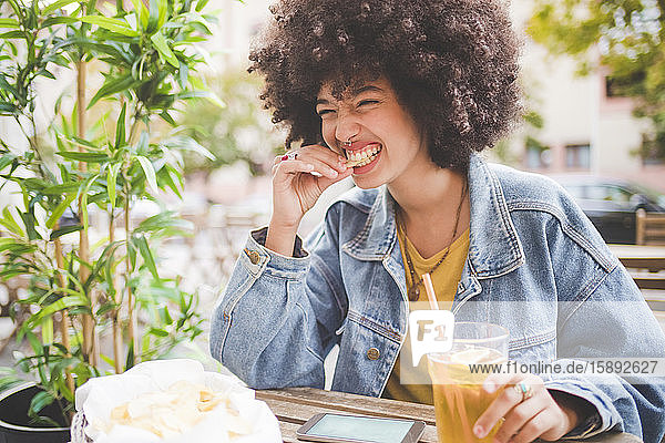 Happy young woman with afro hairdo eating chips at an outdoor cafe in the city