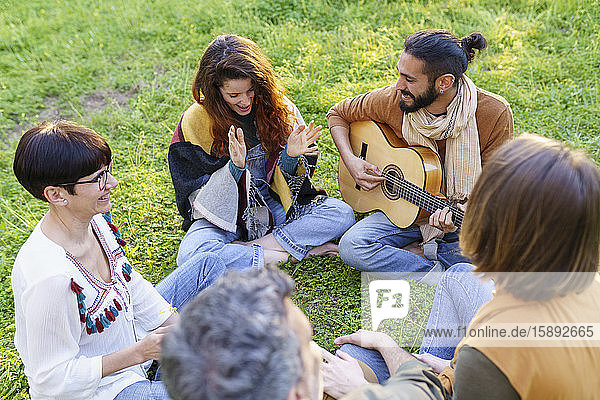 Group of friends playing music with the guitar sitting on the grass in the field
