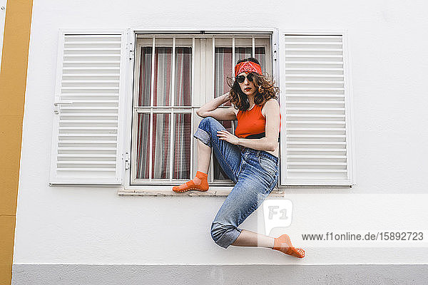 Portrait of young woman wearing headband and sunglasses sitting on window sill outdoors