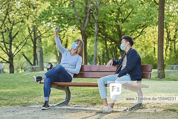 Man and woman wearing a face mask on a park bench in a municipal park  doing selfie  Corona crisis  Regensburg  Bavaria  Germany  Europe