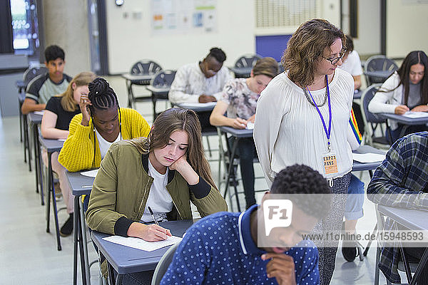 High school teacher supervising students taking exam desks classroom