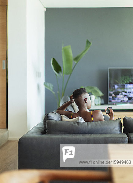 Young woman with remote control watching TV on living room sofa