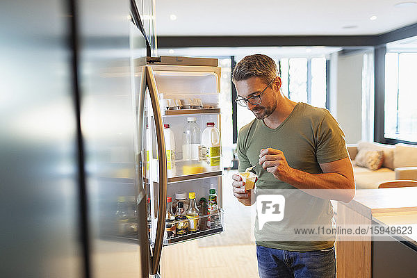 Mann isst Joghurt am offenen Kühlschrank in der Küche