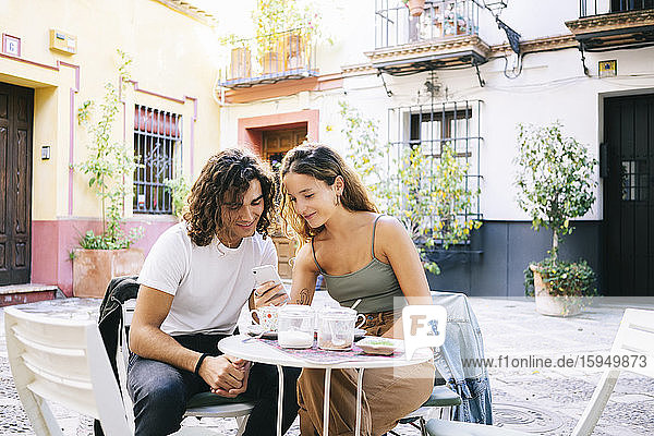 Junge Frau zeigt ihrem Freund ihr Handy  während sie in einem Straßencafé sitzt  Santa Cruz  Sevilla  Spanien