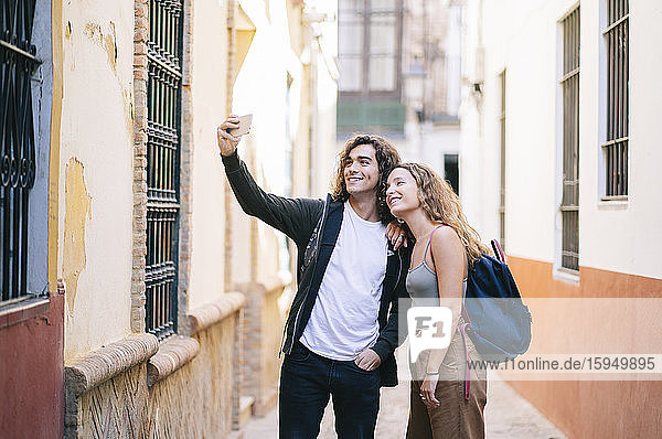 Smiling young couple taking selfie while standing on narrow street at Santa Cruz  Seville  Spain