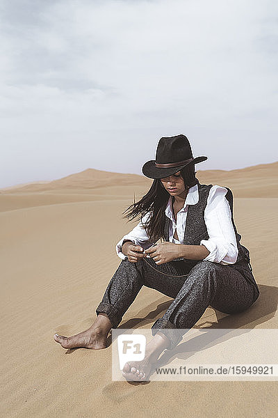 Modische junge Frau sitzt barfuss auf einer Düne und schaut auf eine Taschenuhr  Wüste Merzouga  Marokko