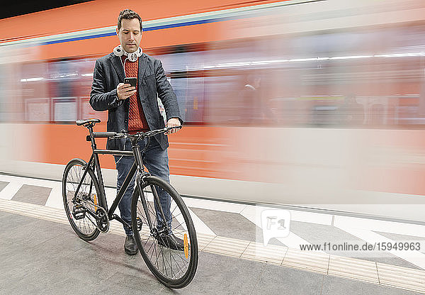 Geschäftsmann mit Fahrrad benutzt Telefon  während er mit der U-Bahn auf dem Bahnsteig geht