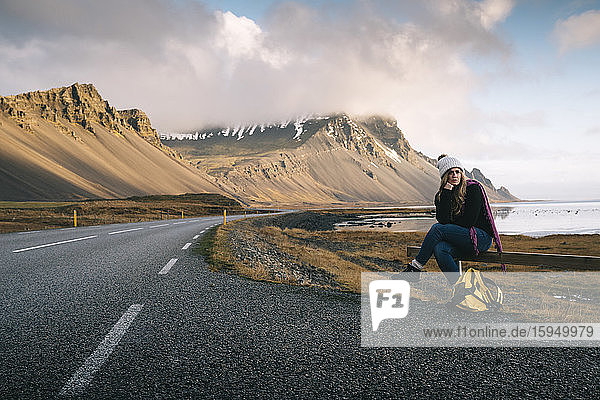 Woman with backpack sitting on railing while waiting at roadside against mountains in Iceland