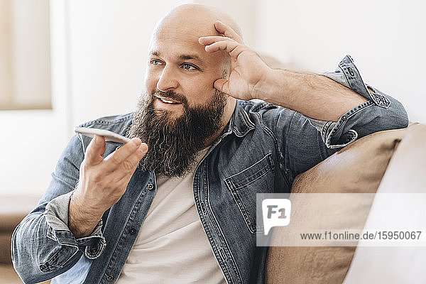 Businessman with shaved head talking over speaker on smart phone while sitting on sofa at home