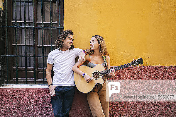 Lächelnder junger Mann steht neben seiner Freundin  die Gitarre spielt  während er an der Wand steht  Santa Cruz  Sevilla  Spanien