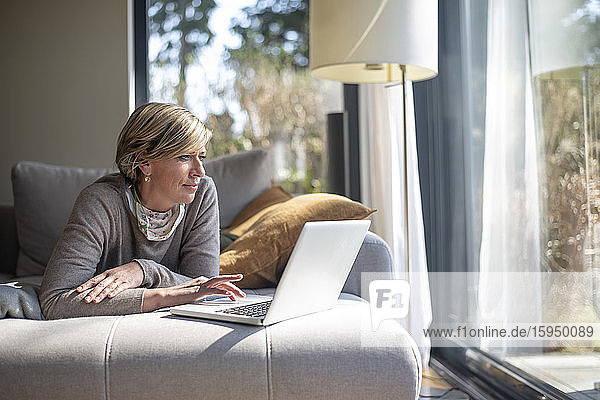 Reife Frau  die während der Arbeit von zu Hause aus einen Laptop benutzt  während sie im Wohnzimmer auf dem Sofa liegt