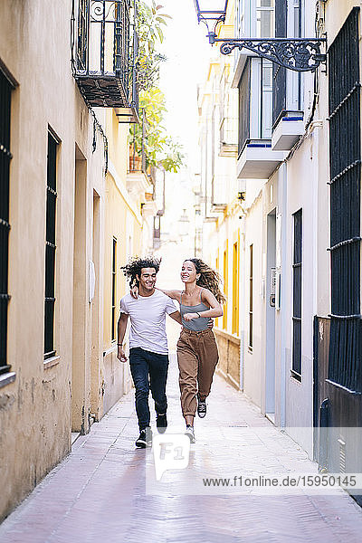 Ein glückliches junges Paar läuft in voller Länge auf einer schmalen Straße in Santa Cruz  Sevilla  Spanien