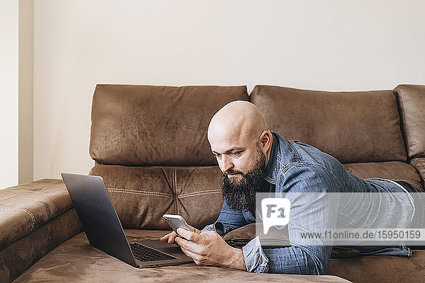 Geschäftsmann mit Laptop  der sein Handy benutzt  während er zu Hause auf dem Sofa liegt