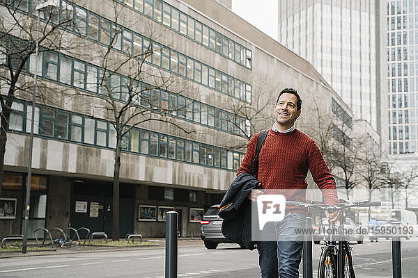 Lächelnder Geschäftsmann schaut weg  während er mit dem Fahrrad auf der Straße in Frankfurt am Main unterwegs ist