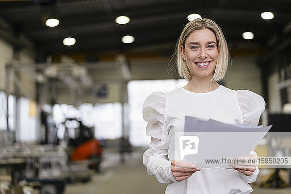 Porträt einer lächelnden jungen Frau  die in einer Fabrik Papiere in der Hand hält
