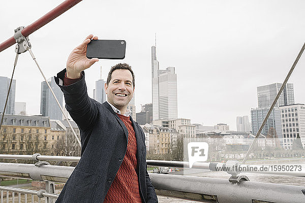 Lächelnder Geschäftsmann  der auf einer Brücke über den Fluss steht und sich mit einem Smartphone selbstständig macht  Frankfurt  Deutschland