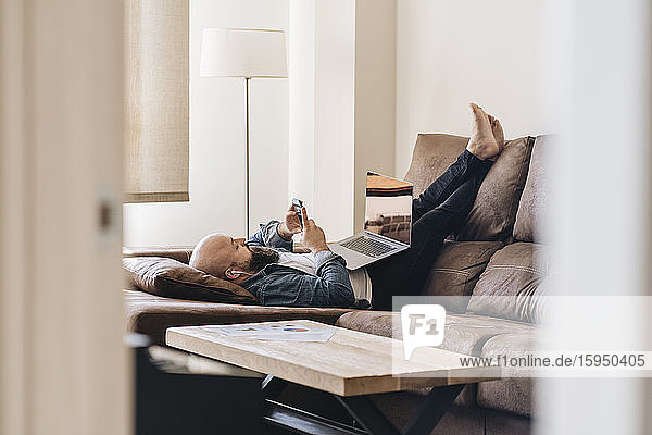 Businessman with laptop listening music through smart phone while lying on sofa at home
