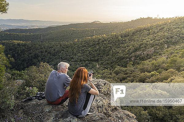 Vater und erwachsene Tochter sitzen auf einem Felsen und genießen die Aussicht bei Sonnenuntergang