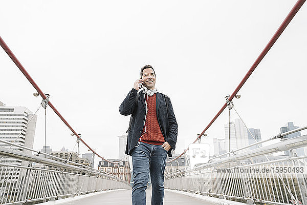 Lächelnder Geschäftsmann spricht am Smartphone  während er auf der Brücke gegen den Himmel in Frankfurt geht