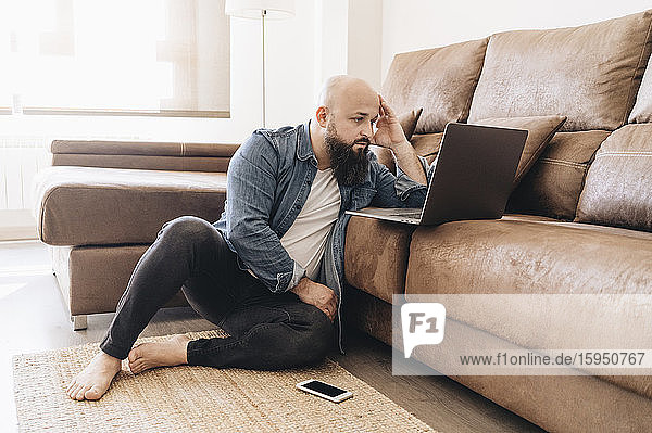 Seriöser Geschäftsmann mit Laptop  während er zu Hause auf dem Boden am Sofa sitzt