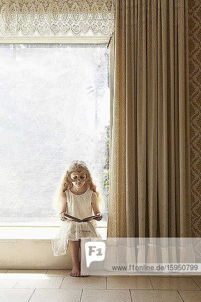 Mädchen sitzt auf der Fensterbank mit Pappbrille und liest Buch
