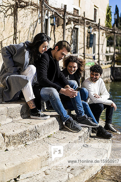 Four friends sitting on stairs at Lake Garda sharing smartphone  Italy
