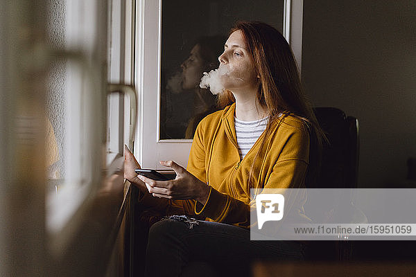 Lächelnde rothaarige Frau mit Smartphone raucht am offenen Fenster