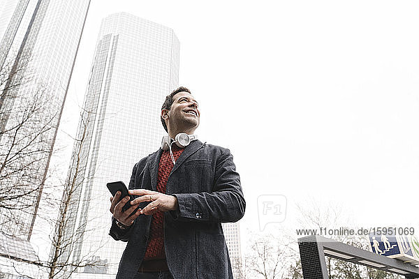 Lächelnder Geschäftsmann mit Smartphone schaut weg  während er in der Stadt steht  Frankfurt  Deutschland