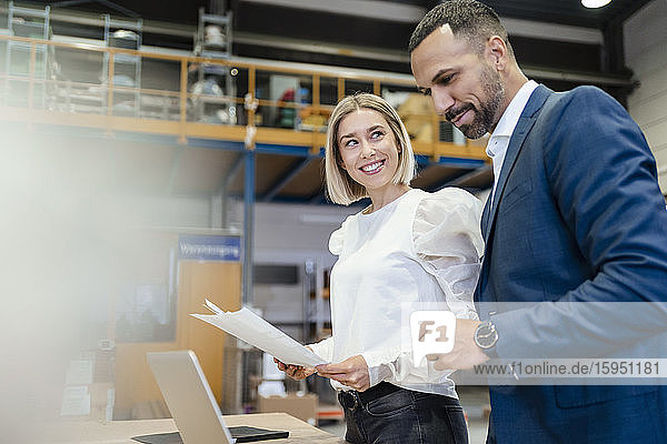 Geschäftsmann und junge Frau mit Papieren und Laptop in einer Fabrik